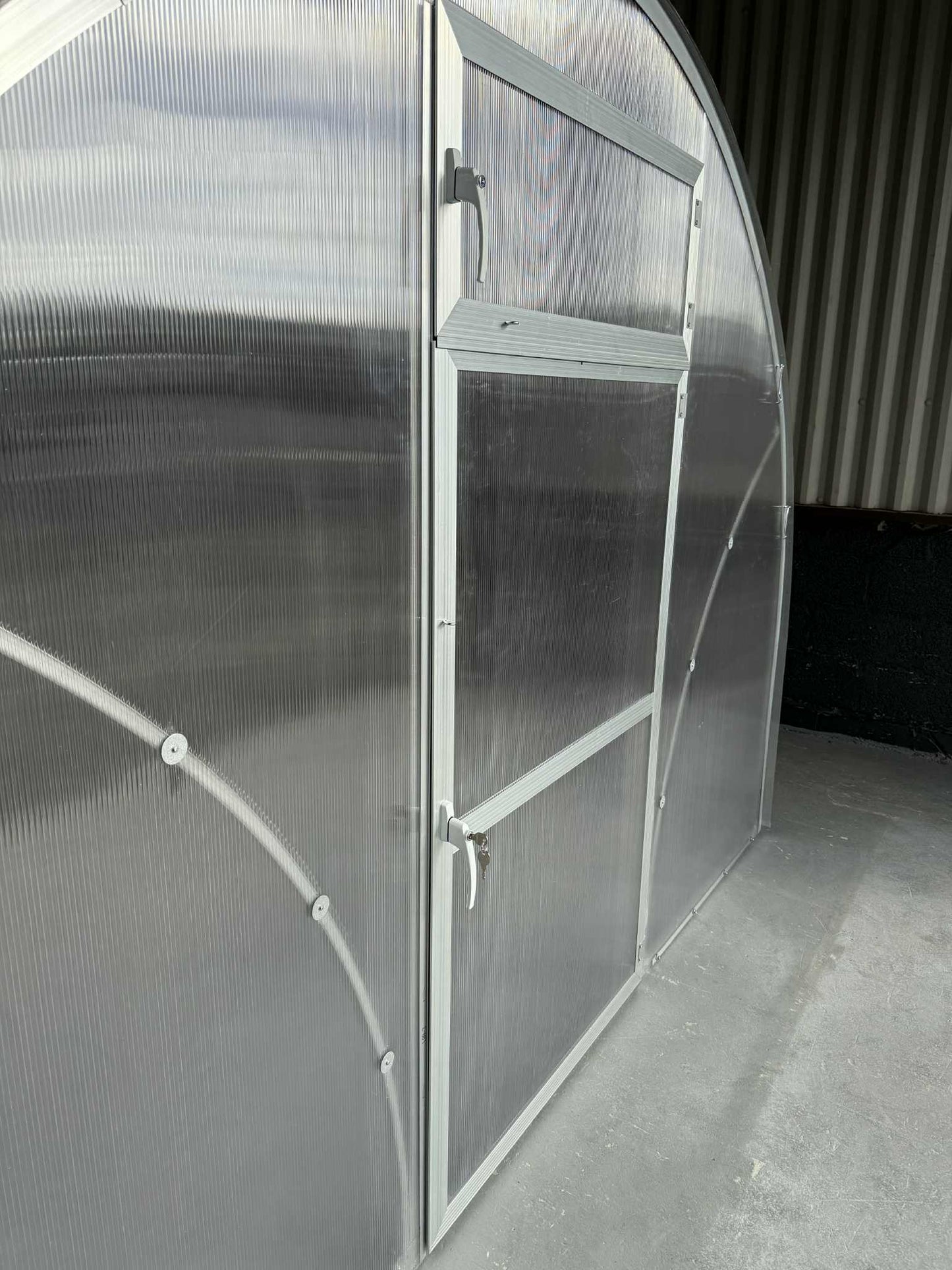 Partial view of a  Sigma 300 (Gen III) greenhouse with a curved glass panel and metal frame.